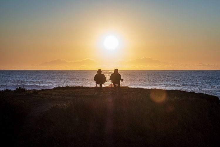 Two people sitting next to the ocean watching the sunrise.