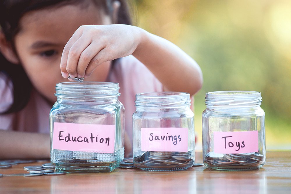 Girl placing coins into jars marked education, savings, and toys.