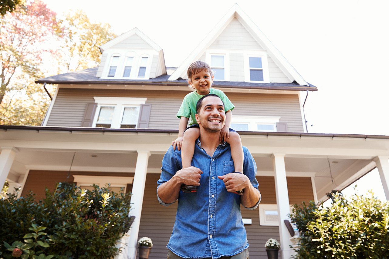 Smmiling Father with son on his shoulders with their house in the background.