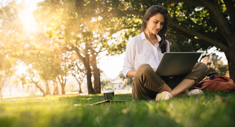 Young Woman with earphones sitting in the grass using a laptop to create a budget.