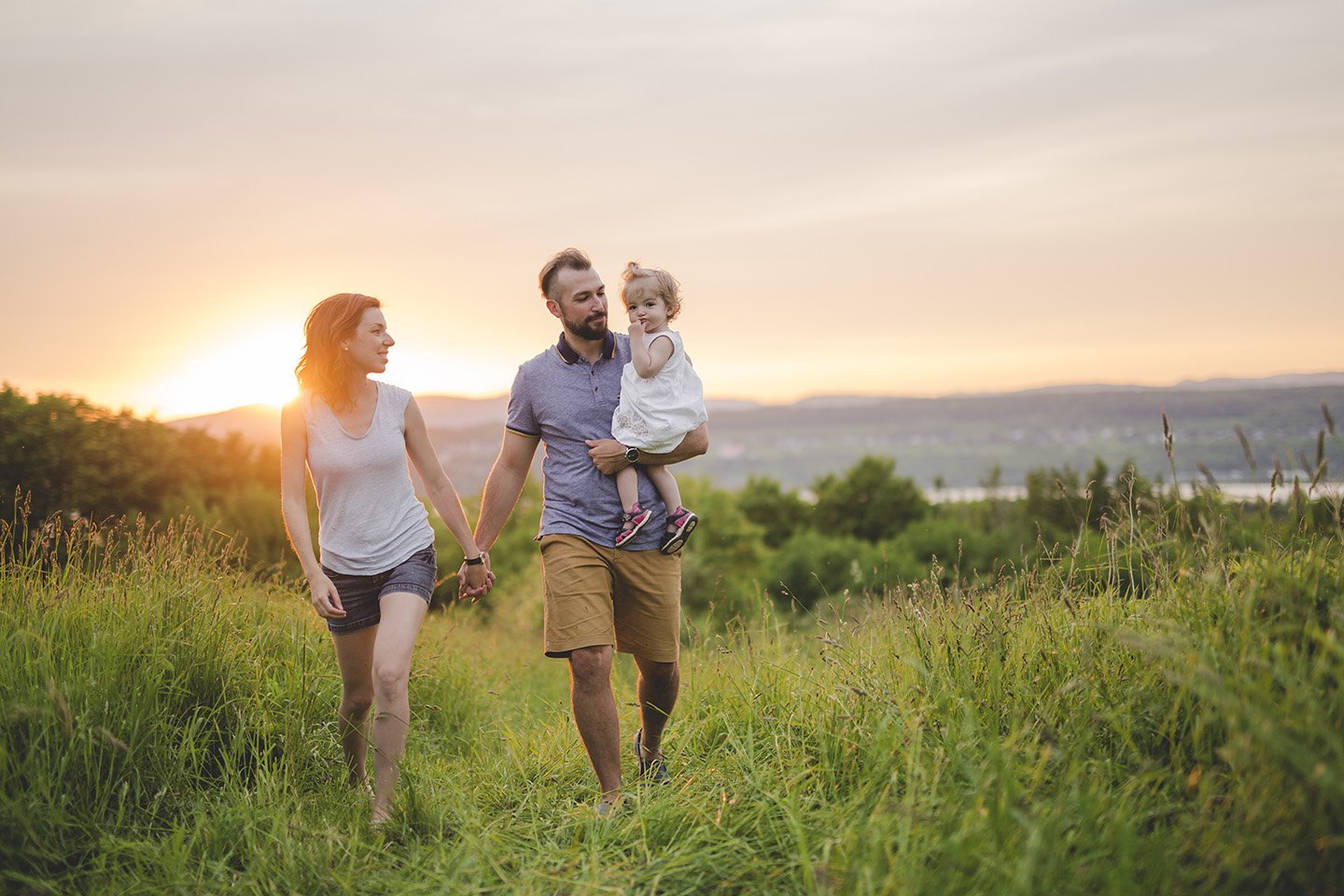 Young couple walking through high grass at sunset holding hands as the father carries their daughter.