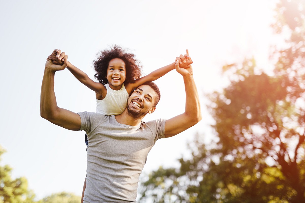 Black father lifting his daughter over his shoulder smiling outdoors.