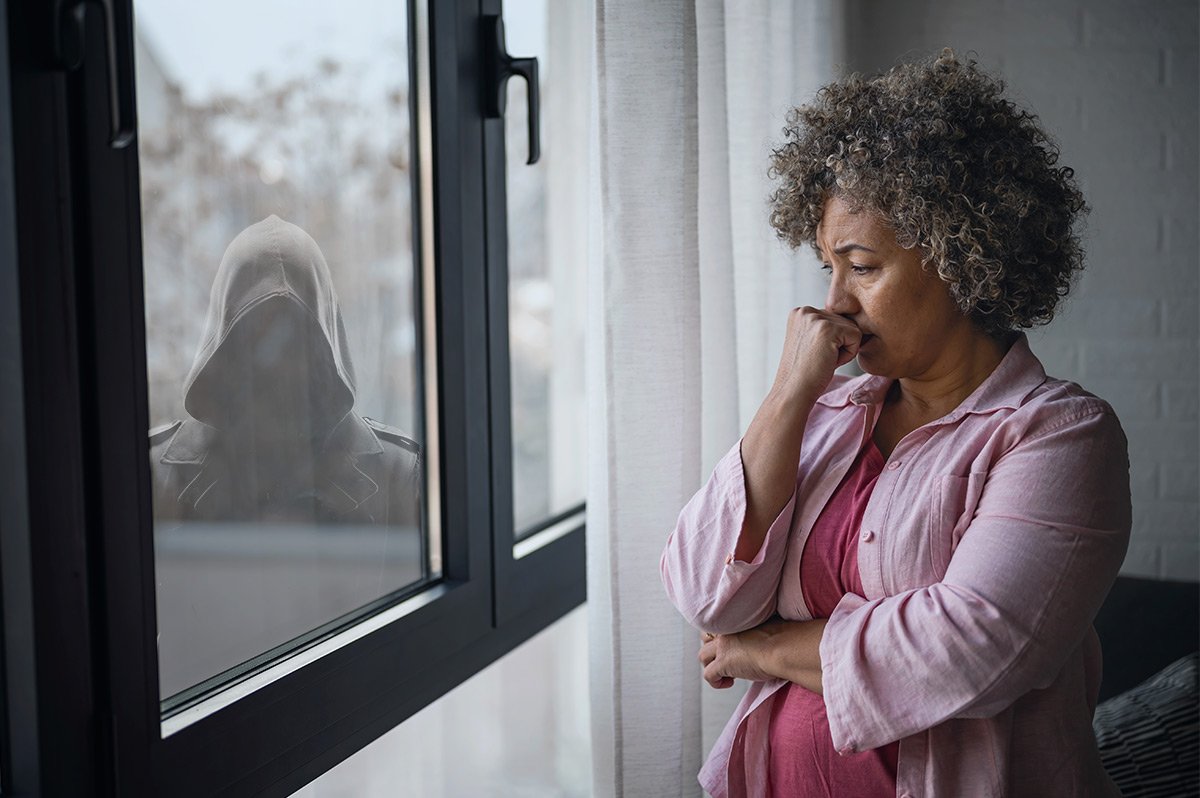 Woman looking down by a window with a hooded man in the background looking in.