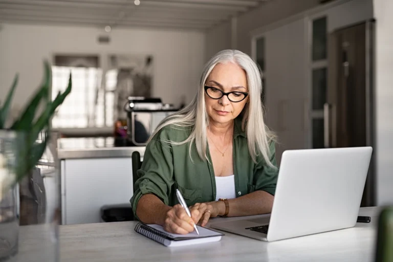 Retired woman sitting at desk with her laptop taking notes with a pen and paper.