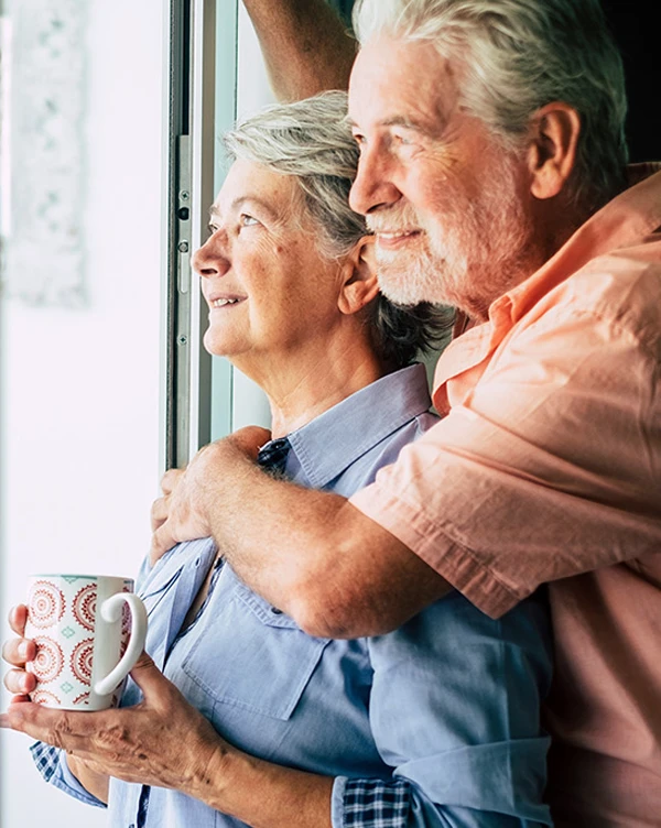 A retired couple embracing and looking out a window.